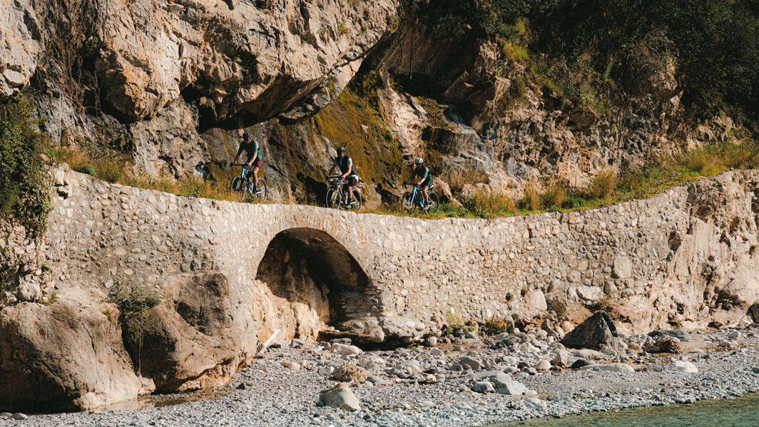 Canyon Gravel Bike Buying Guide – Choosing the Right Bike for Every Adventure Group of cyclists riding Canyon gravel bikes across a stone bridge carved into a rocky mountainside, highlighting the bikes' off-road capability and adventure readiness.